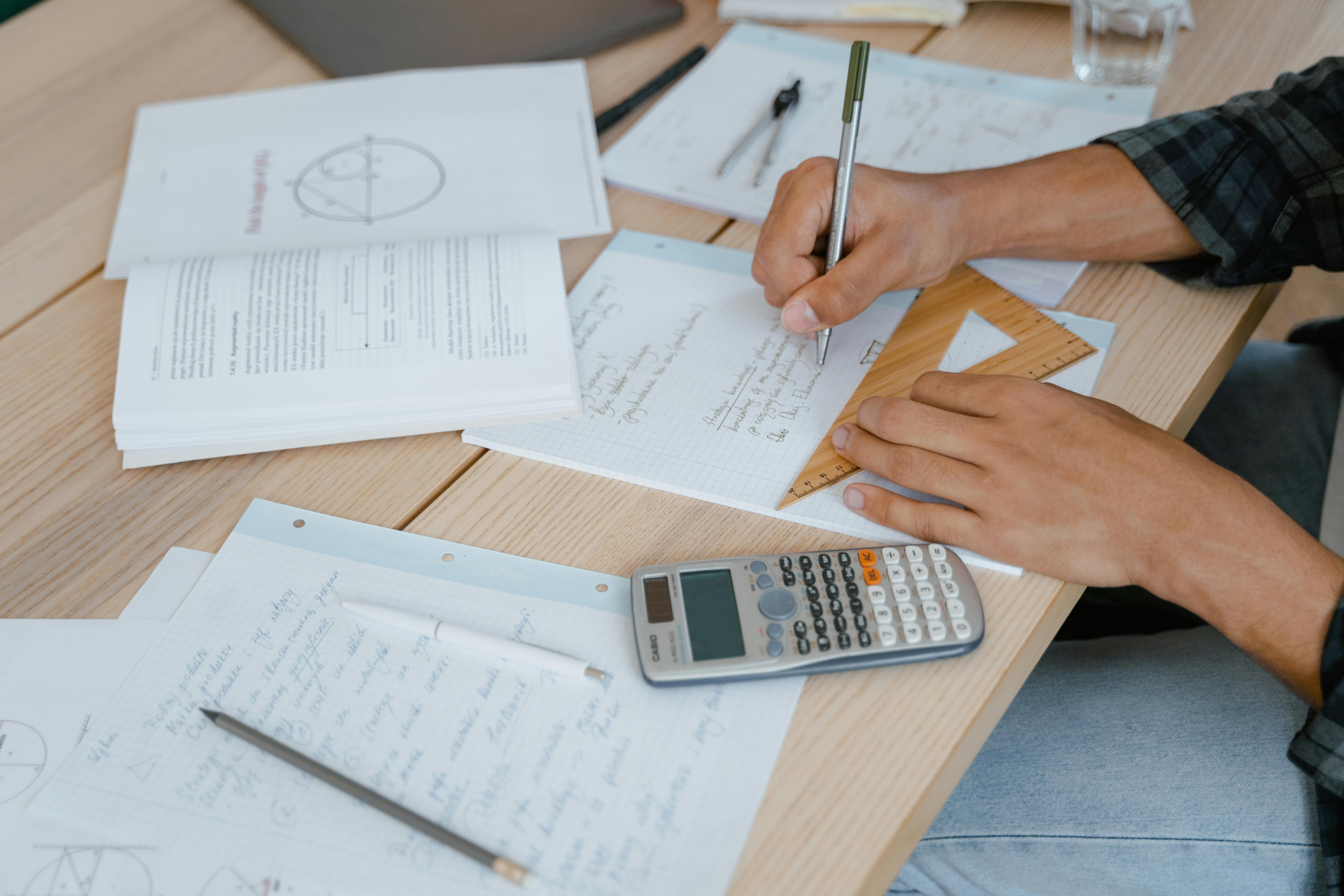 Student hands using wooden triangle ruler and scientific calculator while solving mathematical problems on paper with notebook